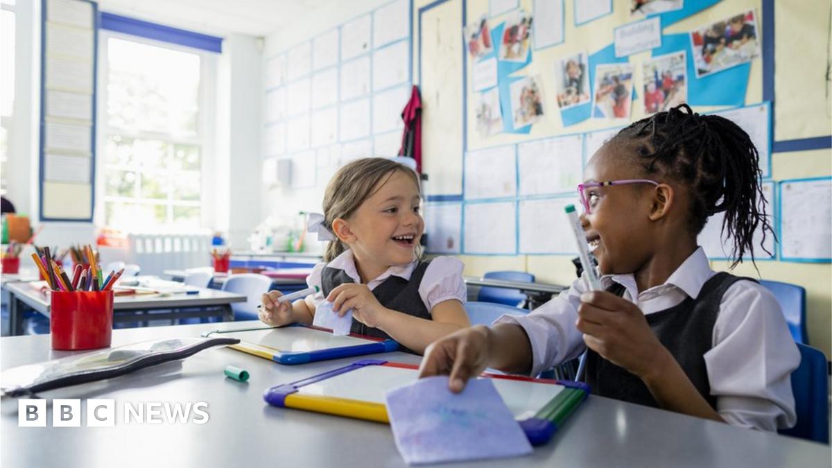 Two children smile at each other while sat at their desks in a primary classroom.